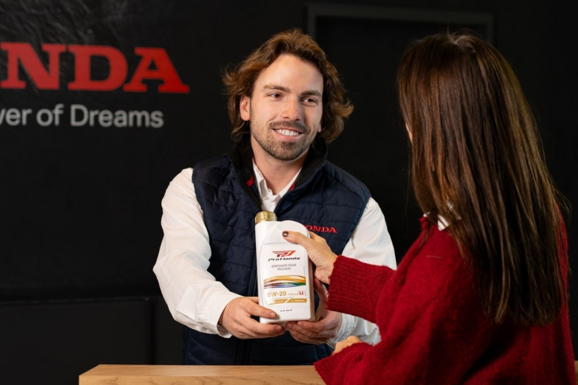 A Honda employee in a navy vest hands a bottle of Honda engine oil to a customer in front of a black wall with the Honda logo and slogan \"Power of Dreams\". The employee is smiling as he presents the product.